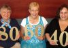 A photo of Letha Dawson Scanzoni, Linda Bieze, and Alena Ruggerio. Each is holding a gold paper number of their age. Letha holds an 80, Linda holds a 60, and Alena holds a 40.